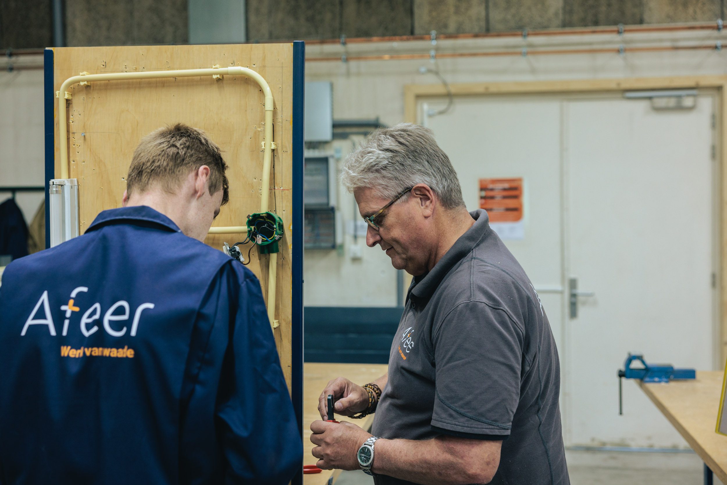 Een medewerker is in het TechniekLab met bedrading bezig, terwijl een instructeur van Afeer toekijkt